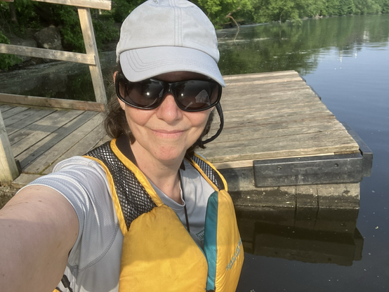 A woman with large sunglasses and sage green ball cap, life jacket and grey T-shirt in front of wooden dock