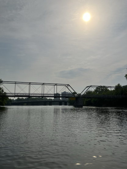 A metal and wood bridge spans the small river with a sign saying construction ahead, waterway closed and the sun shining above through hazy skies