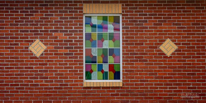 A 12-pane window with multi-colored patterns of glass in a brick wall accented with lighter bricks in a diamond shape on either side of the window.