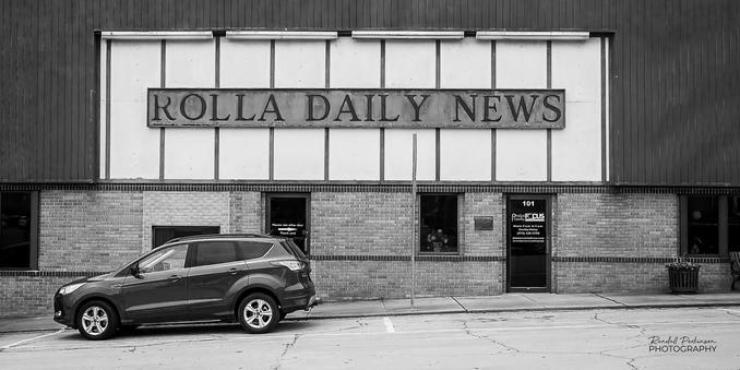 Black and white image of a brick building with a large sign that reads "Rolla Daily News".  A small SUV is parked on the street in front of the building.