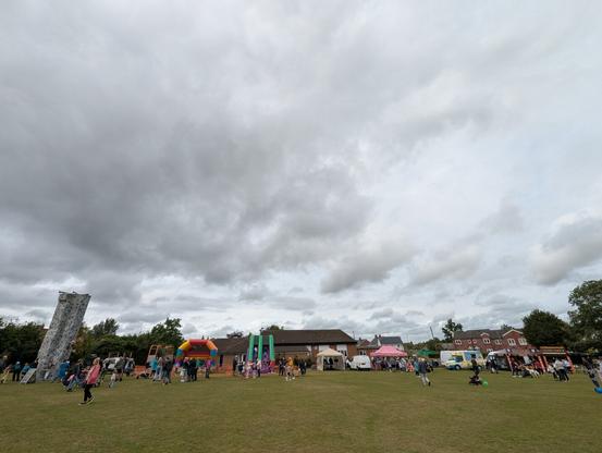 A wide angle view of a village fete in progress complete with climbing wall, bouncy castles, DJ booth, ice cream van and food stalls. Above, a very dynamic and threatening sky is beginning to clear.
