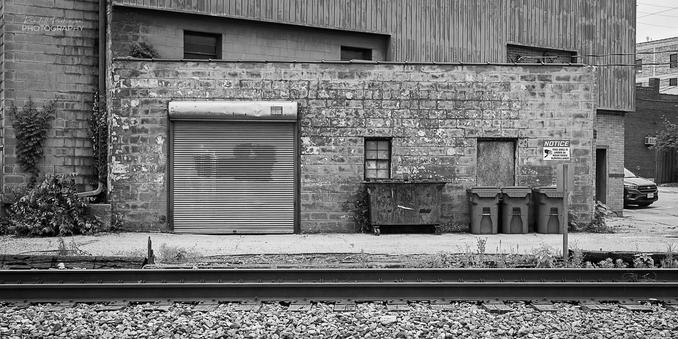 A one-story brick building with a garage and two windows opens onto a narrow drive between the building and a set of railroad tracks. A dumpster and three trash receptacles sit adjacent to the building.