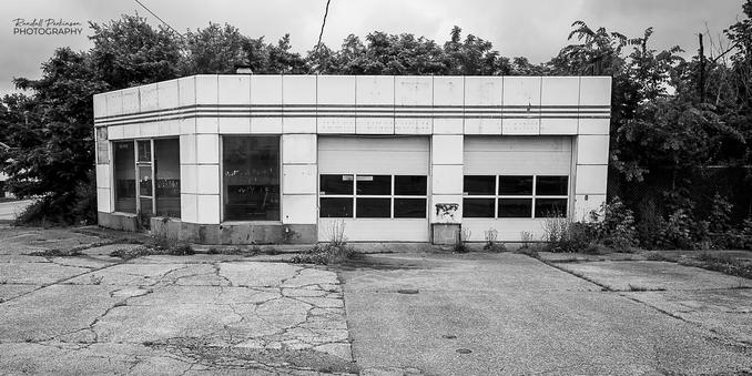 An old, abandoned service station with an office area on the left and two garage doors on the right.  Weeds are growing out of cracks in the concrete pavement surrounding the building.