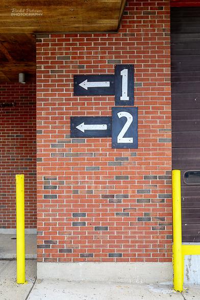 A brick wall has signs with white numbers on black background that read "1" and "2".  Each of these signs has an arrow sign adjacent to it pointing to the left.