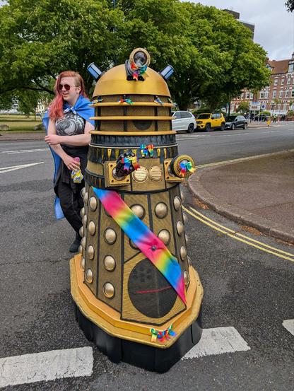 A gold Dalek from Doctor Who is standing on a street. It has rainbow decorations. The wide, curving street beyond had trees offering shade to the cars parked by it.