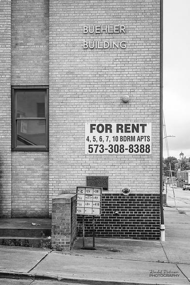 An outside view of the Buehler Building, an apartment building just blocks from Missouri S&T.  A sign on the building reads, "FOR RENT - 4, 5, 6, 7, 10 BDRM APTS - 573-308-8388".  A freestanding mailbox containing 12 separate boxes stands in front of the building.