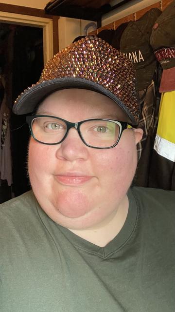 A selfie of me in our mudroom. I’m wearing an orange/golden rhinestone cap, green glasses and a green shirt. Behind me some more caps and coats are visible