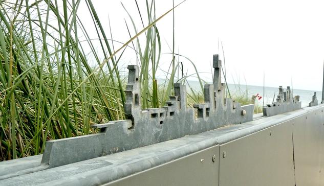 Metal cutout of war ships on a low wall, just behind are beach grasses and in the far distance a small canadian flag, sand, and light blue water. The day is overcast and the sky is grey.