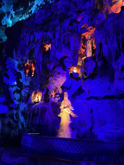 A wall in a limestone cave, bathed in blue light with small statues illuminated in red and orange.