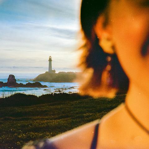 Au tout premier plan, flou, l'épaule droite et la moitié droite du visage d'une jeune femme de face. Derrière elle, net, on aperçoit un paysage côtier sans doute au soleil couchant : une prairie qui descend vers la mer, quelques rochers au large, un phare de l'autre côté d'une petite baie.