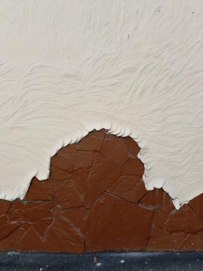 The photograph depicts a brown stone wall with a white plaster-like material covering most of its surface. At the bottom of the photograph, a small section of dark asphalt is visible, which is sprinkled with white powder that is probably a result of the crumbling plaster.