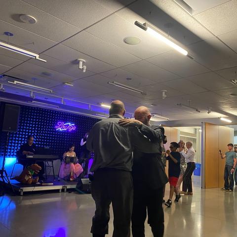 In the foreground, a man in a suit dances wirh a person in grey shirt/pants and tall black boots. In the background, more couples dance. A keyboardist, bandeonist, and violinist are visible onstage.