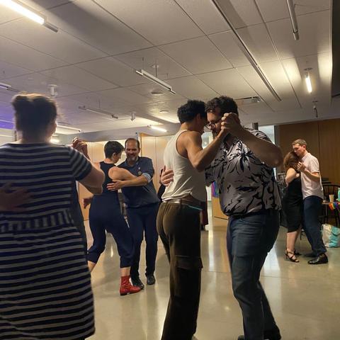 Four dancing couples. On the left, there’s the back of a woman in a striped dress. Somewhat centered is a curly-haired person in a white tank top and brown pants dancing with a bearded person in a floral shirt. In the background, two of our organizers, Georgia, in red boots, leads José, and another couple dances.