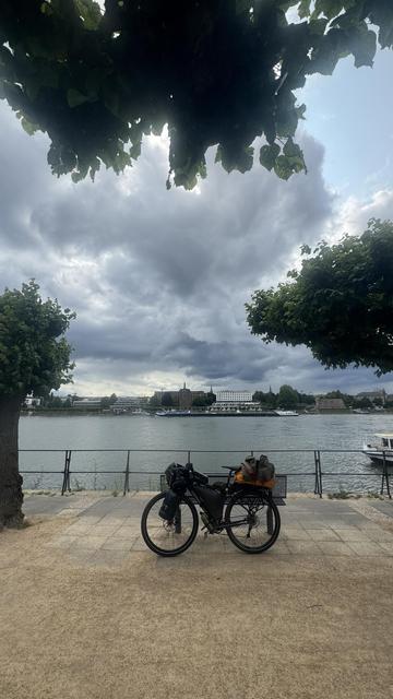 Ein Fahrrad mit Taschen, die an einem Flussufer geparkt sind, umrahmt von Bäumen und einem bedeckten Himmel. Die Szene zeigt ruhiges Wasser und abgelegene Gebäude entlang der Küste.