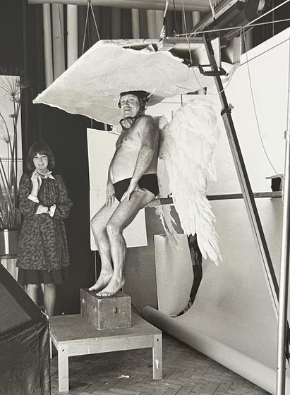 Side view of the shot showing the Angel balancing on a seat with the ground perched on his head. A studio assistant looks on smiling.