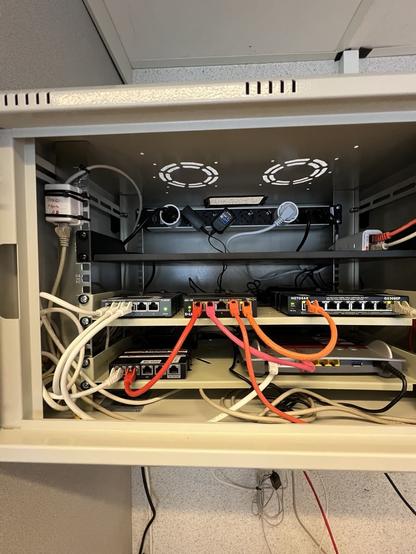 Man sieht einen kleinen Netzwerkschrank mit 3 Einlegeböden. Auf dem untersten Boden liegt eine Fritz.Box und ein ERX. Auf dem mittleren Boden liegen zwei 5-Port Switche und ein 8-Port-Switch.