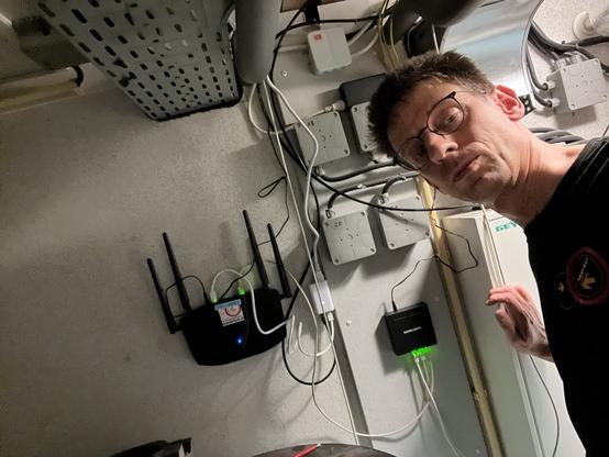 PharmaJoe steht nach dem Materialaustausch in einem von sechs Technikräumen. Im Hintergrund zu sehen: 1 Totolink Router, 1 POE Netzteil, 1 5-Port-POE-Switch. Im Hintergrund viele Strom-Verteiler-Dosen und eine Metal-Kabel-Brücke.