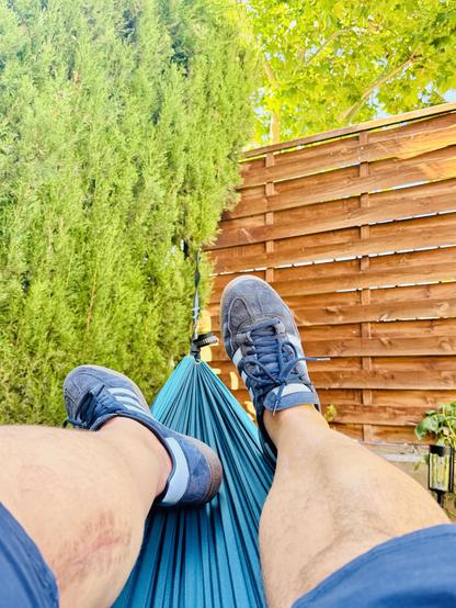 Fotografía en color que muestra a una persona descansando en una hamaca azul, con los pies calzados en zapatillas deportivas azules. Al fondo se observa una valla de madera, un ciprés y un árbol de hojas verdes. La luz es intensa, con predominancia de tonos verdes, marrones y azulados.

Color photograph showing a person resting in a blue hammock, with their feet wearing blue sneakers. In the background, there is a wooden fence, a cypress tree, and another leafy green tree. The lighting is bright, with predominant green, brown, and blue tones.