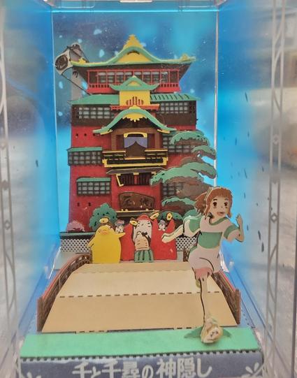 A completed paper model kit showing the scene when Chihiro leaves the bath house in the movie Spirited Away. All the spirits are waving at her as she runs off with a smile on her face.