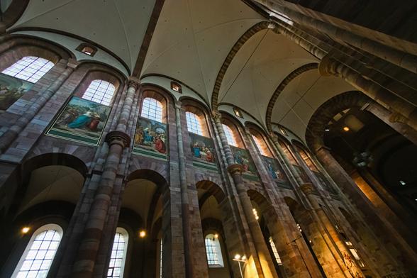 Ein schräger Blick die Wand einer hohen Kirche entlang Richtung Decke. Wir sehen unten Fenster, darüber Bilder an der Wand, und oben nochmal eine Reihe Fenster. Rechts scheint die Sonne orange herein.