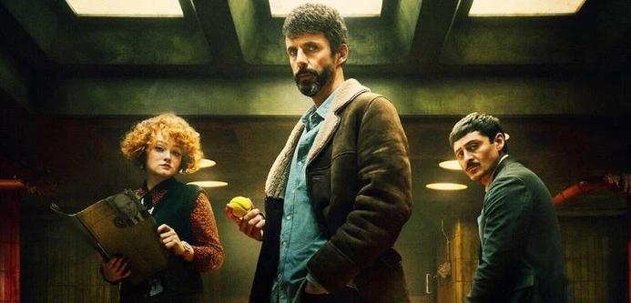 Three characters stand in a dimly lit room. A man at the center, holding a yellow object, has a serious expression. To his left, a woman with curly hair holds a clipboard, while to his right, another man appears focused. The
