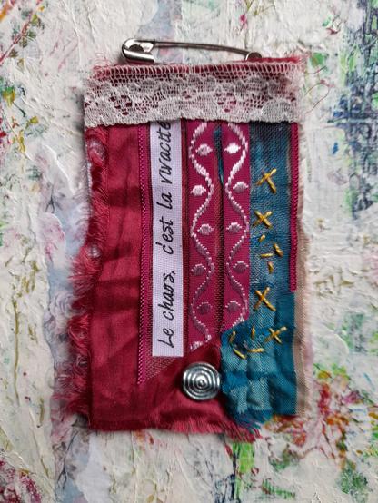 Le chaos c'est la vivacité = Chaos is liveliness. Collage with red and green silk, paper, lace and slow stitching in yellow. A silver bead with a spiral.