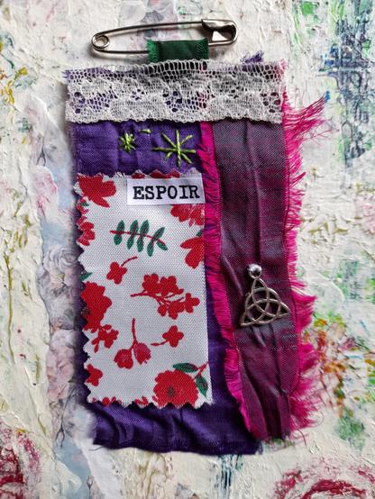 Floral fabric in red and green, purple silk, lace and slow stitching. A silver triskell and the word éspoir for hope.