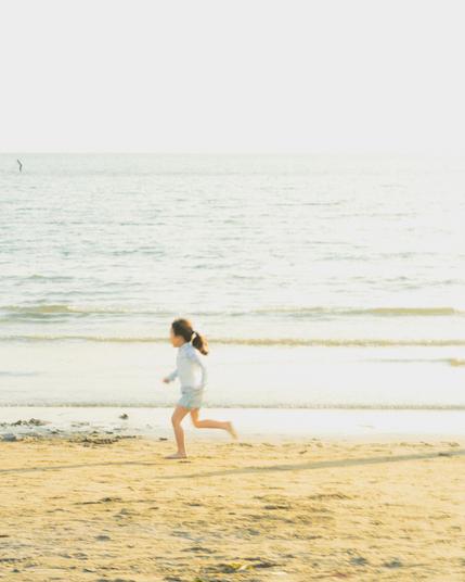 A young girl with a ponytail runs along a sandy beach near the water's edge. The sea is calm with gentle waves and a bright, hazy sky above. A solitary figure is visible in the distant water. The scene is bright and sunny, casting soft shadows on the sand.