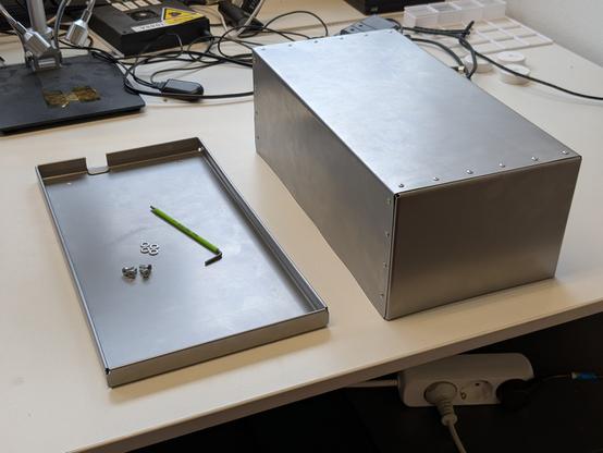 A picture of a sheet metal box sitting on a table. On the left, there is a shallow tray with a small notch in the back.  On the right a riveted box is shown, which can be placed onto the tray.