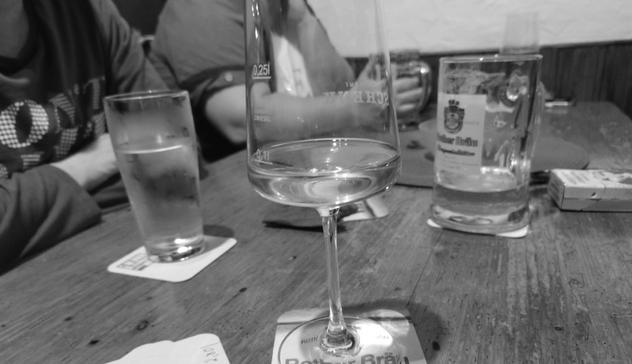 Ein Schwarzweiß-Foto zeigt einen Holztisch in einer Bar oder einem Restaurant mit verschiedenen Getränken: Im Vordergrund steht ein Weinglas mit Weißwein auf einem runden Bierdeckel, dahinter befinden sich ein hohes Glas mit hellem Bier, eine Wasserflasche und weitere Gläser. Im Hintergrund sind verschwommen Menschen zu sehen, die an dem Tisch sitzen.