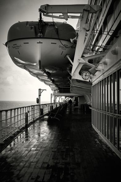 A black-and-white image showing the promenade deck of the Queen Mary 2. The deck is wet from rain, and the ship is against a backdrop of the North Atlantic Ocean. Two people are walking along the deck under life boats. There's a clock and railing along the side.