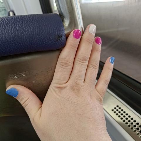 A picture of a hand against the seatback of a train seat. The nails are painted in the trans flag colors: blue, pink, white, pink, blue.