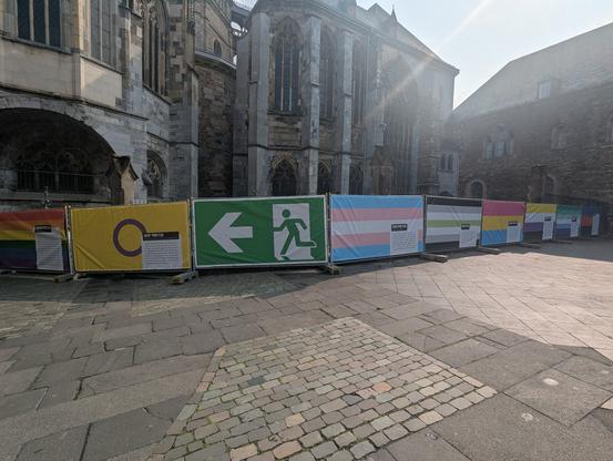 A bunch of big flags covering a temporary fence. From left to right:

6 striped pride flag,
Inter pride flag,
ISO7010 emergency exit sign,
Trans flag,
Agender flag,
Pan flag,
Non-binary flag,
Gay men pride,
Polyamorous flag,