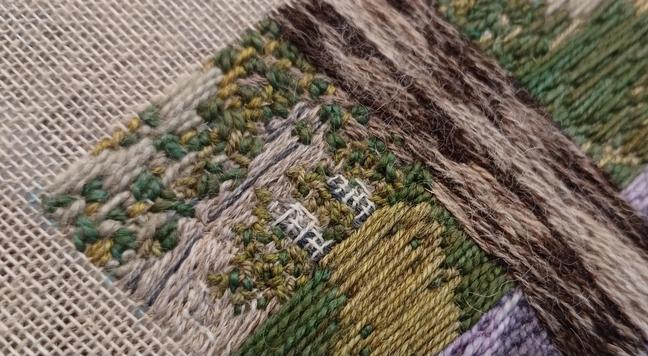 Corner of a stitched artwork depicting the corner of a stately home, part hidden by leaves and behind a hedge.