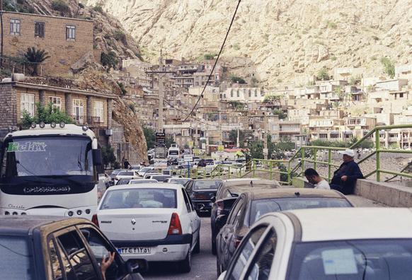 A busy street in a mountainous village with heavy traffic, brick houses built on the hillside, and two men sitting by a railing in the foreground.
