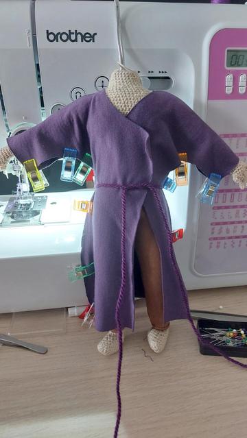 A crocheted doll standing in front of a white and pink sewing machine. The doll is crocheted out of light beige yarn and is wearing camel brown fitted trousers and a purple wizard robe that overlaps on the front. The sides are pinned together with fabric clips.
