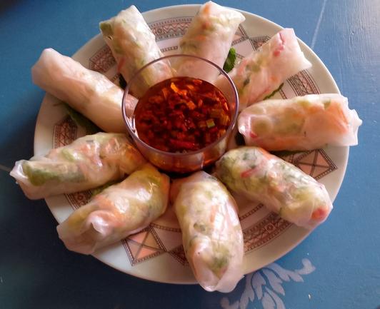 Un cuenco con una salsa de color rojo intenso. A su alrededor nueve rollitos hechos con papel de arroz rellenos de verduras.