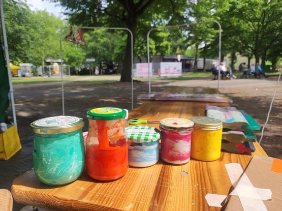Jars with color inside on a table in the sunlight, in the background there are trees and benches with people sitting on them