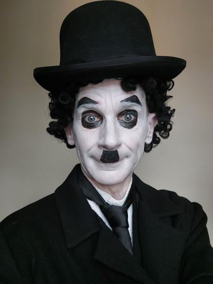 Selfie au format vertical de votre serviteur costumé en Charlot, le personnage inventé en 1914 par Charles Chaplin, chapeau melon noir, perruque noire, visage maquillé de blanc et de noir avec la moustache, chemise blanche, cravate noir, gilet et redingote noirs.