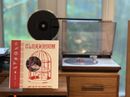 Cloakroom - Last Leg of the Human Table LP cover.

A white cover with an OBI strip with symbols. Inside the cover is a transparent overlay of a birdcage with the title - I've put it outside so it's visible.

The damnable glitter LP plays on a vintage, restored Bang & Olufsen Beogram 4000 linear-tracking turntable to the right. The tonearm looks dismissively at the stupid glitter specks.