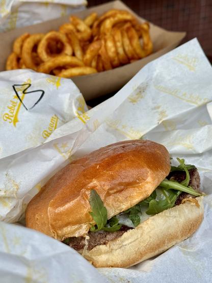 A close-up of a juicy burger with a golden bun, fresh greens, and condiments is in the foreground, surrounded by white paper. In the background, there is a serving of curled, golden-brown fries in a brown tray.