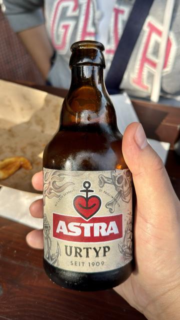 A hand is holding a bottle of Astra Urtyp beer, featuring a label with a heart and nautical theme. The background shows some blurred food items and clothing.