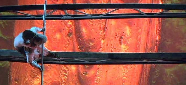 The image shows two individuals wearing white outfits clinging to a vertical pole. They are positioned on metallic beams that extend horizontally across the scene. The background features a vivid, fiery orange surface that resembles molten lava or an intensely lit wall, creating a stark contrast with the characters in the foreground. The composition emphasizes a tense moment in a dramatic environment.