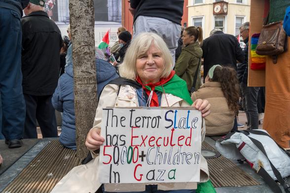 A protest scene on Grand Parade, Cork, showing a person sitting on the ground among a crowd, holding a handmade sign that reads: "The terror State has Executed 50,000+ Children In Gaza." The person is draped in a flag of Palestine, and other demonstrators with placards and flags are visible in the background.