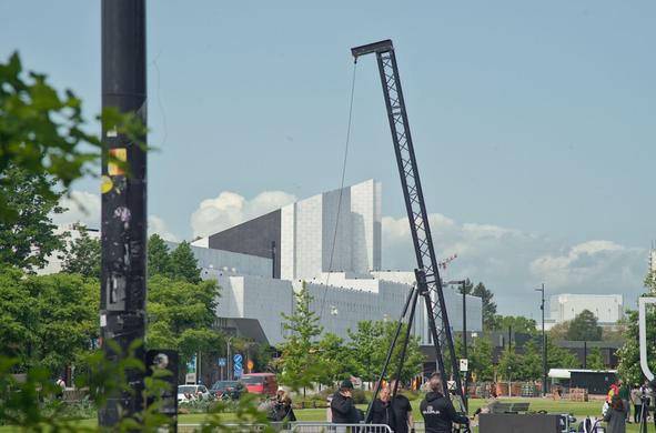 A multi-layered photograph of the eastern side of Finlandia Hall, an angular and white building. On the foreground out-of-focus foliage, on the mid-ground a small crane operated by a crew of workers preparing a stage and passers by.

The photo makes it seem like the photographer was hiding when the shot was taken.