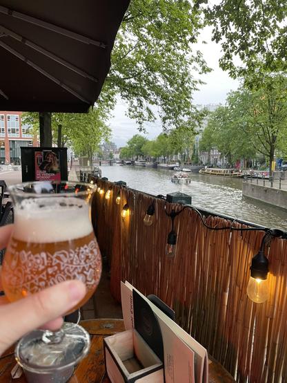 A aomewhat artistic photo of a hand holding a glass of beer with an Amsterdam canal in the background. There is a bamboo fence decorated with pendant lights between the camera and canal.