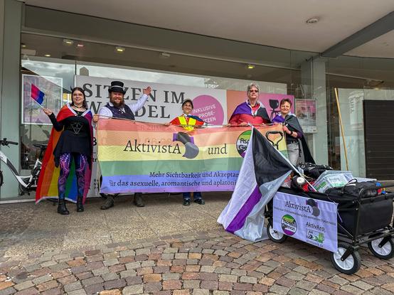 Fünf Menschen posieren hinter einem bunten Banner. Die Personen haben sich verschiedene Pride-Flaggn umgehängt oder sind passend kostümiert. Auf dem Banner steht "AktivistA und Aspec*German - Mehr Sichtbarkeit und Akzeptanz für das asexuelle, aromantische und aplatonische Spektrum." Im Vordergrund steht ein Bollerwagen, aus dem eine Demi-Ace-Flagge ragt. Ein kleines Textilbaner macht Werbung für AktivistA, aromantik.de und Aspec*German.