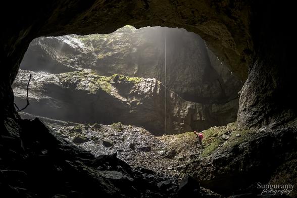 A large passage window to an open air pit entrance to a cave system. We can see a rope hanging as a line of white just right of center. There is greenery in the pit from moss and ferns. A lone caver traverses across wearing a bright red pack and helmet - they are tiny and show the scale of this cave.