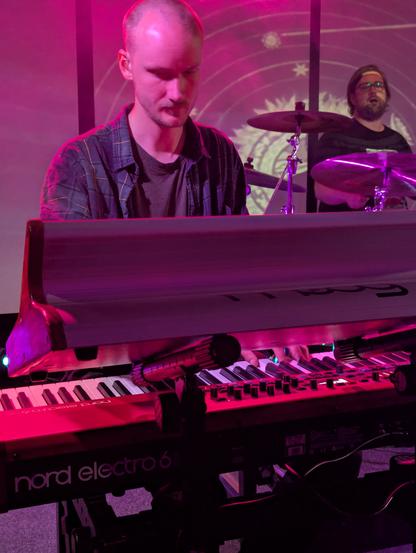 Foto från dimmig livescen, musiker syns bakom keyboard och trummor.
