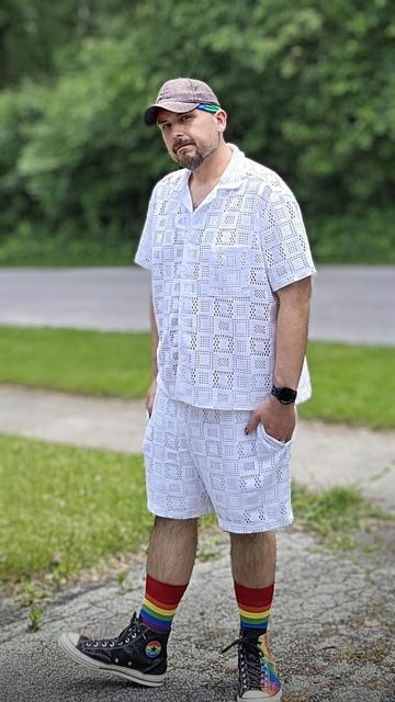 Matt B stands outdoors on a sunny day, wearing a matching white crochet-style short-sleeve shirt and shorts set. His outfit is semi-sheer, with a geometric square pattern throughout. He accessorizes with a black baseball cap featuring a rainbow bandana underneath, small  earrings, and a black smartwatch. He's also wearing rainbow-striped socks and black high-top Converse shoes that feature a rainbow star patch. Behind him is a quiet residential street and a lush green backdrop of trees and grass.